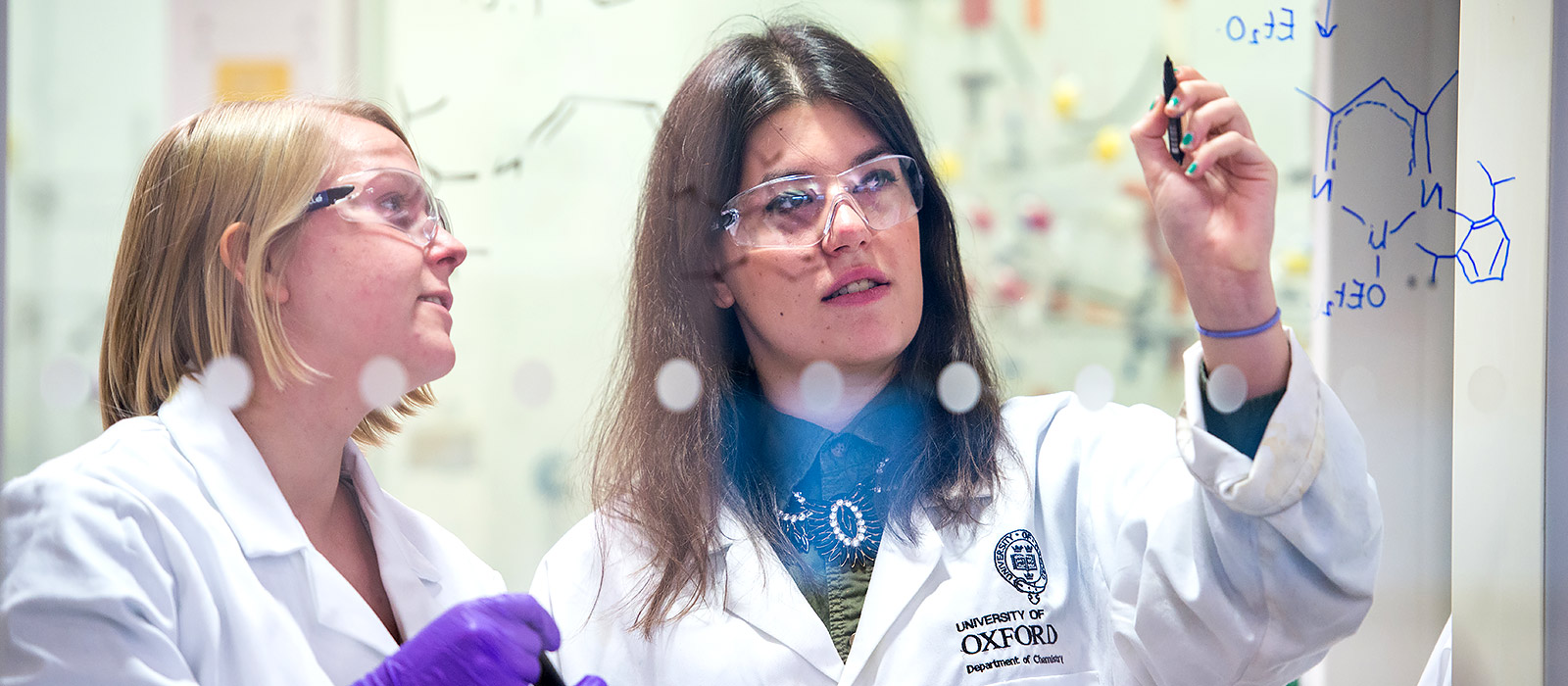 Two students wearing lab coats write out formulas on a windowpane in the chemistry lab