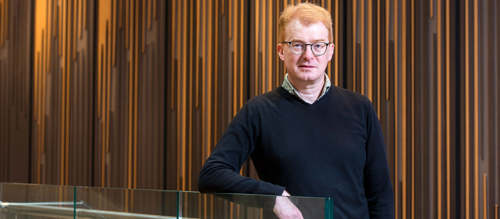 Professor Matt Higgins stands in front of a richly toned panelled wall